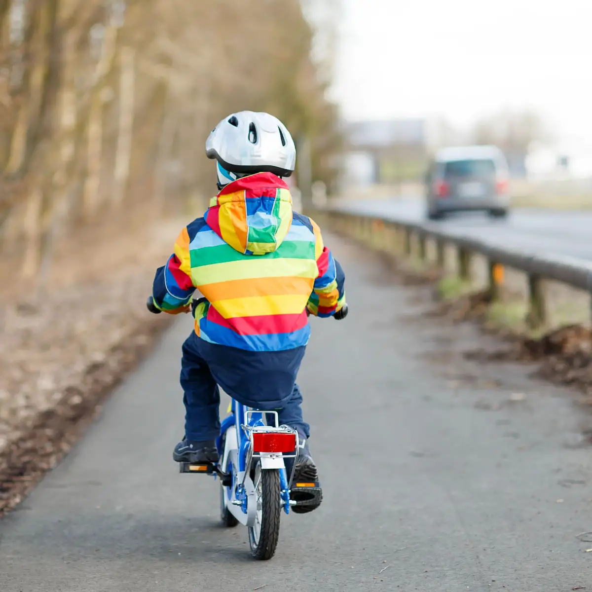 Little kid boy in safety helmet and colorful raincoat riding his first bike and having fun on cold day, outdoors. From back, street with cars.