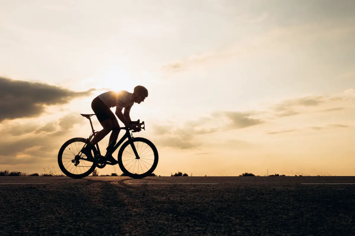 Side view of active sportsman in protective helmet riding bike among nature during sunset. Muscular guy in activewear cycling on paved road.